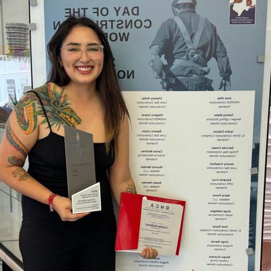 A woman holds an award and certificate in front of a photo backdrop.